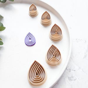 Puede incluir: Un juego de seis cortadores de galletas en forma de lágrima, con cinco de color marrón claro y uno de color morado. Los cortadores están dispuestos en círculo sobre un plato blanco.