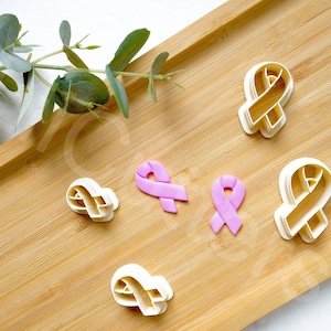 Puede incluir: Un conjunto de seis cortadores de galletas de cinta rosa sobre una superficie de madera. Los cortadores son de varios tamaños y formas, y algunos están decorados con fondant rosa.