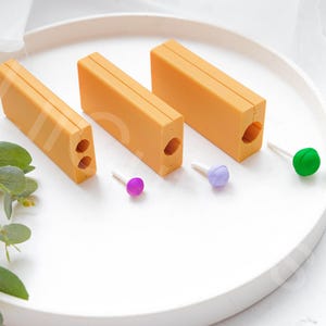 Pode incluir: Três blocos retangulares de cor laranja com furos de tamanhos diferentes. Os blocos estão dispostos do maior para o menor. Há três pequenos palitos de pirulito coloridos ao lado dos blocos.