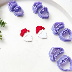 Pode incluir: Um conjunto de seis cortadores de biscoitos de plástico roxo em forma de Papai Noel. Dois cortadores são do rosto do Papai Noel com um chapéu vermelho e uma barba branca, e quatro cortadores são do chapéu do Papai Noel.