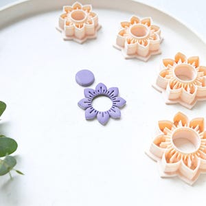 May include: Five flower-shaped cookie cutters on a white plate. Four are peach-colored and one is purple. The cutters are arranged in a circle around a small purple circle of clay.