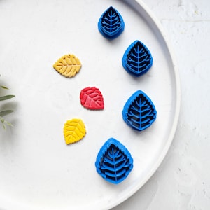 May include: A set of five blue leaf-shaped cookie cutters on a white plate. The cutters are different sizes and have a detailed design. There are also three small leaf-shaped cookies in yellow, red, and yellow.