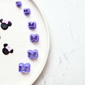 Peut inclure: Une assiette blanche avec sept emporte-pièces en forme de nœud papillon en plastique violet de différentes tailles. Trois oreilles de Mickey Mouse en fondant noir avec des nœuds roses sont sur le côté gauche de l'assiette.