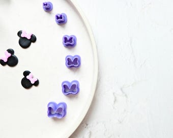May include: A white plate with seven purple plastic bow-shaped cookie cutters of varying sizes. Three black fondant Mickey Mouse ears with pink bows are on the left side of the plate.