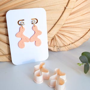 Könnte beinhalten: Pfirsichfarbene, abstrakte Hängeohrringe mit strukturierter Oberfläche, präsentiert auf einer weißen Karte. Die Ohrringe haben einen kleinen, funkelnden Akzent oben. Passende Ohrringausstecher sind ebenfalls sichtbar.