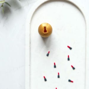 May include: A gold sphere with a small red lipstick cutout on a white surface with several red lipsticks scattered around it.