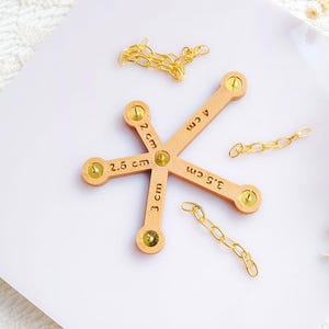 Puede incluir: Un calibrador de cadena de madera de color claro con medidas en centímetros, rodeado de cadenas doradas. El calibrador tiene cinco brazos, cada uno etiquetado con una longitud diferente, de 2 cm a 4 cm. Las cadenas son de diferentes longitudes.