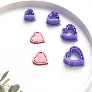 Pode incluir: Um conjunto de cinco cortadores de biscoitos em forma de coração roxo em uma superfície branca. Também há dois biscoitos em forma de coração rosa na superfície.