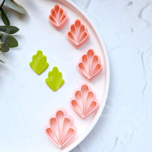 May include: A set of pink and green plastic cookie cutters shaped like seashells and leaves. The cutters are on a white plate.