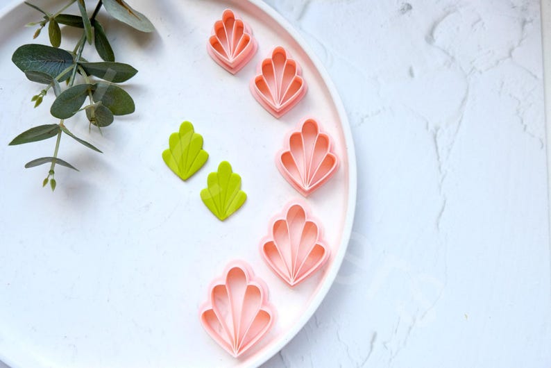 May include: A set of pink and green plastic cookie cutters shaped like seashells and leaves. The cutters are on a white plate.