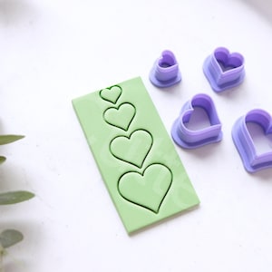 Può includere: Un set di cinque tagliapasta a forma di cuore di diverse dimensioni, con un tappetino da forno in silicone verde che presenta cinque sagome a forma di cuore.