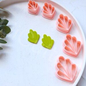 May include: A set of pink and green plastic cookie cutters shaped like seashells and leaves. The cutters are on a white plate.