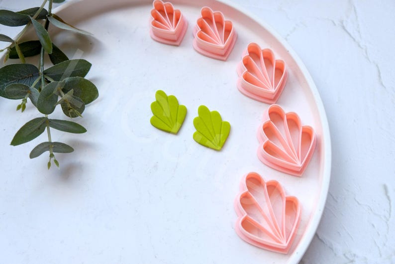 May include: A set of pink and green plastic cookie cutters shaped like seashells and leaves. The cutters are on a white plate.