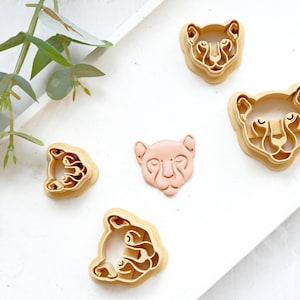 Peut inclure: Cinq emporte-pièces en forme de visages d'animaux. Les emporte-pièces sont en plastique et de différentes nuances de brun et de rose. Les emporte-pièces sont disposés sur une surface blanche.