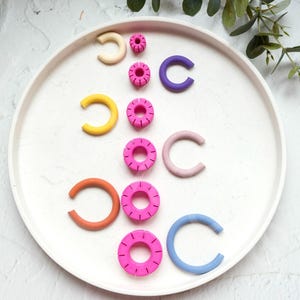 May include: A set of colourful, handmade polymer clay earrings. The earrings are in the shape of a circle and a half-circle. The colours are pink, yellow, orange, blue, and purple.