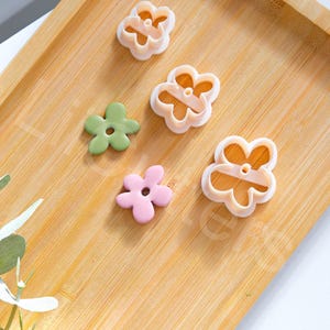 Può includere: Quattro stampini per biscotti a forma di fiore in plastica bianca su una superficie di legno. Ci sono anche tre pezzi di pasta di zucchero a forma di fiore in rosa, verde e rosa chiaro.
