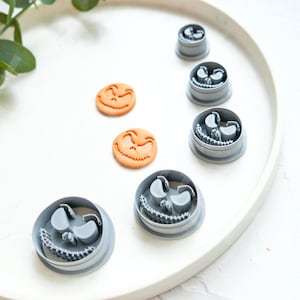 Puede incluir: Un juego de seis cortadores de galletas de plástico gris con forma de la cara de Jack Skellington de la película Pesadilla antes de Navidad. Los cortadores están dispuestos en círculo sobre un plato blanco. También hay dos galletas naranjas en el plato.