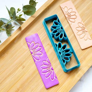 Op de afbeelding: Drie koekjesvormen in de vorm van madeliefjes op een houten oppervlak. Eén vorm is blauw, één is roze en één is perzikkleurig.