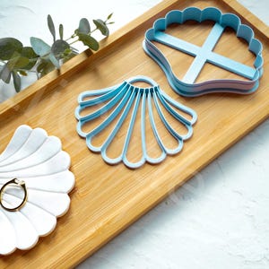 May include: Three blue seashell-shaped cookie cutters on a wooden tray. The cookie cutters are different sizes and have different designs.