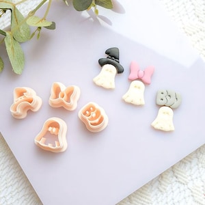 Può includere: Una collezione di formine per biscotti e decorazioni a tema Halloween. Le formine hanno la forma di fantasmi, fiocchi e zucche. Le decorazioni includono fantasmi con cappelli e fiocchi. I colori sono rosa chiaro, nero e bianco.