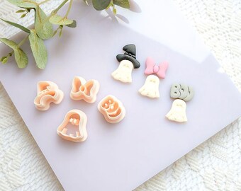 May include: A collection of Halloween-themed cookie cutters and decorations. The cutters are in the shape of ghosts, bows, and pumpkins. The decorations include ghosts with hats and bows. The colors are light pink, black, and white.