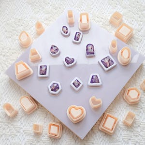 May include: A collection of light peach-colored polymer clay cutters in various shapes, including hearts, squares, and teardrops. Some cutters contain purple and clear resin pieces. The cutters are arranged on a light purple surface.