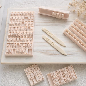May include: A set of light beige plastic letter and number stamps for clay or dough, including a large grid tray, two smaller grid trays, a handle, and three long pointed markers with the embossed words THYME and BASIL clearly visible.