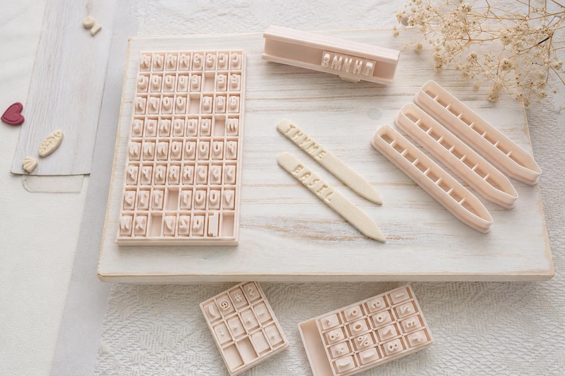 May include: A set of light beige plastic letter and number stamps for clay or dough, including a large grid tray, two smaller grid trays, a handle, and three long pointed markers with the embossed words THYME and BASIL clearly visible.