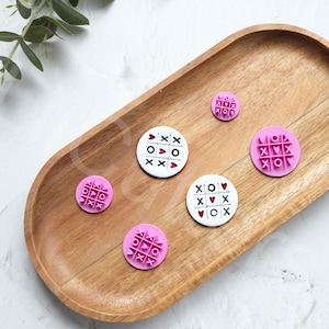 May include: A set of six pink and white round wooden stamps with a tic-tac-toe design and hearts. The stamps are on a wooden tray.