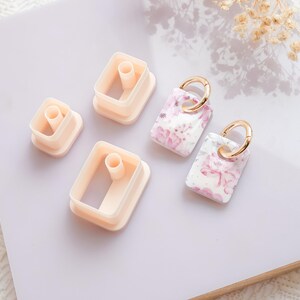 Puede incluir: Imagen de suministros de artesan&iacute;a y pendientes terminados. Hay varios cortadores rectangulares de color melocot&oacute;n claro y dos pendientes rectangulares con aros dorados. Los pendientes tienen una base blanca con estampados florales rosas.