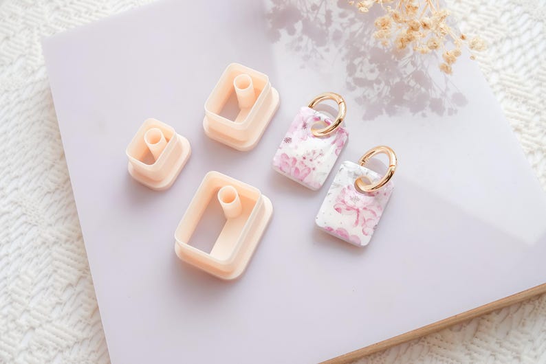 Puede incluir: Imagen de suministros de artesan&iacute;a y pendientes terminados. Hay varios cortadores rectangulares de color melocot&oacute;n claro y dos pendientes rectangulares con aros dorados. Los pendientes tienen una base blanca con estampados florales rosas.