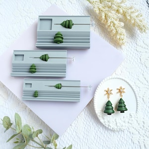 Könnte beinhalten: Weihnachtsschmuck und Dekorationen werden präsentiert. Das Bild zeigt drei graue, rechteckige Präsentationsstücke mit grünen Baumakzenten sowie ein Paar grüne Baumohrringe mit goldenen Schneeflockenaufsätzen. Die Artikel sind auf einer weißen Spitzentischdecke angeordnet.