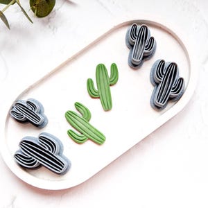 Puede incluir: Un juego de seis cortadores de galletas con forma de cactus, tres son verdes y tres son grises. Los cortadores están dispuestos en un plato ovalado blanco.