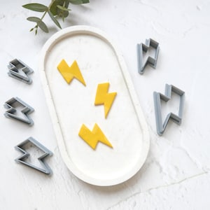 Può includere: Un set di cinque tagliabiscotti a forma di fulmine grigi e quattro biscotti a forma di fulmine gialli su una superficie bianca.