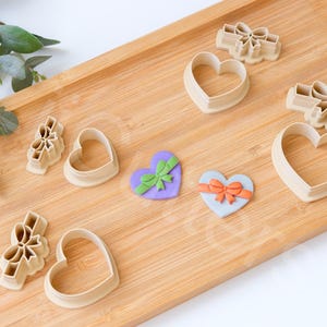 May include: A collection of heart and bow-shaped cookie cutters and decorated hearts on a wooden tray. The cutters are beige, and the hearts are purple and blue, decorated with green and orange bows. Greenery is in the background.