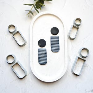 May include: Two pairs of black and white striped clay earrings with black circles on top. The earrings are displayed on a white oval plate with four grey plastic molds shaped like people.