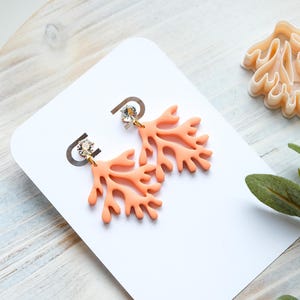 May include: Coral-shaped earrings in a light orange hue, adorned with a small, sparkling crystal at the top. The earrings are displayed on a white card, with a matching coral-shaped object nearby. The earrings are a unique accessory.