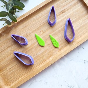 May include: Five purple plastic leaf-shaped cookie cutters and two green leaf-shaped fondant pieces on a wooden surface.