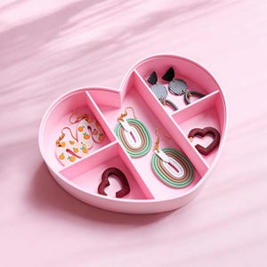 May include: A pink heart-shaped jewellery box with six compartments. The box contains various pairs of earrings, including a pair of orange letter earrings, a pair of green and white striped earrings, a pair of brown and white striped earrings, and a pair of black and white geometric earrings.