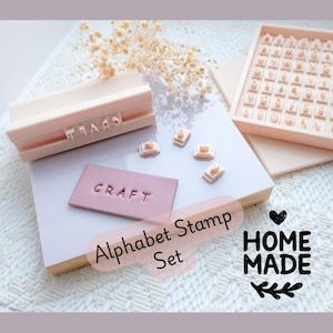 May include: A pink alphabet stamp set with the word "CRAFT" stamped on a pink rectangle. The set includes a tray of letters and a stamp holder. The image also includes the text "Alphabet Stamp Set" and "HOME MADE".