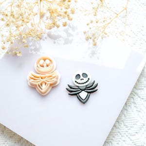 Peut inclure: Deux emporte-pièces, l'un beige et l'autre noir et blanc, sont présentés sur une surface blanche. L'emporte-pièce beige a un design en couches. L'emporte-pièce noir et blanc a la forme d'un personnage avec un visage souriant.