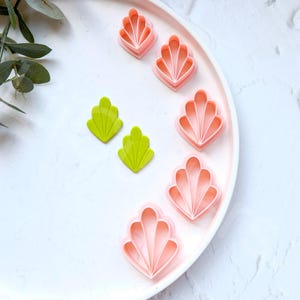 May include: A set of pink and green plastic cookie cutters shaped like seashells. The cutters are arranged on a white plate.