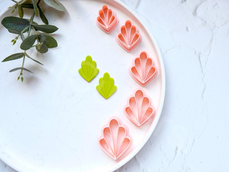 May include: A set of pink and green plastic cookie cutters shaped like seashells. The cutters are arranged on a white plate.