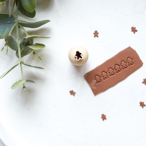 Könnte beinhalten: Ein weißer Teller mit einem braunen Ton-Nudelholz und einem Lebkuchenmann-Ausstecher. Das Nudelholz hat eine Reihe von Lebkuchenmann-Ausschnitten. Auf dem Teller liegen auch mehrere einzelne Lebkuchenmann-Ausschnitte.