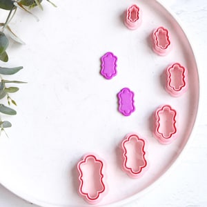 May include: A set of pink and purple plastic cookie cutters in the shape of a rectangle with rounded corners. The cutters are arranged in a semi-circle on a white plate.