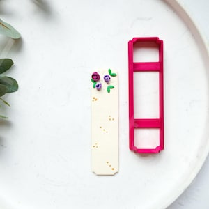 May include: A white bookmark with a pink and purple floral design and gold glitter. A pink plastic bookmark cutter is also shown.