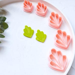 May include: A set of pink and green plastic cookie cutters shaped like seashells and leaves. The cutters are arranged on a white plate.