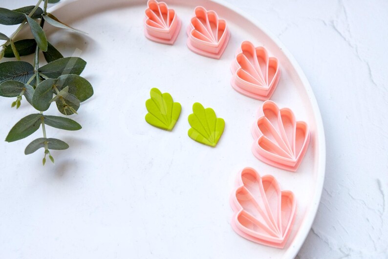 May include: A set of pink and green plastic cookie cutters shaped like seashells and leaves. The cutters are arranged on a white plate.