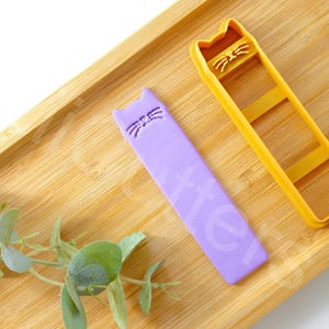 Peut inclure: Deux emportes-pièces en forme de chat, l'un violet et l'autre jaune, sur une surface en bois. L'emporte-pièce violet est un simple contour de tête de chat, tandis que l'emporte-pièce jaune a un design plus détaillé avec un visage et un corps de chat.