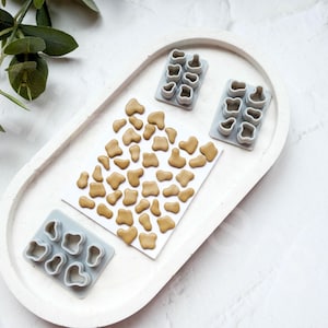 Könnte beinhalten: Ein weißes ovales Tablett mit drei grauen Silikonformen zum Herstellen von Keksen oder Fondant. Die Formen sind mit kleinen, braunen, unregelmäßig geformten Fondantstücken gefüllt.